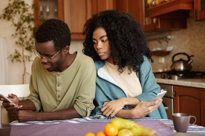 Portrait of sneaky and jealous young African wife spying on her husband, looking over shoulder, trying to see what he reading on his mobile phone. People, relationships and modern technology