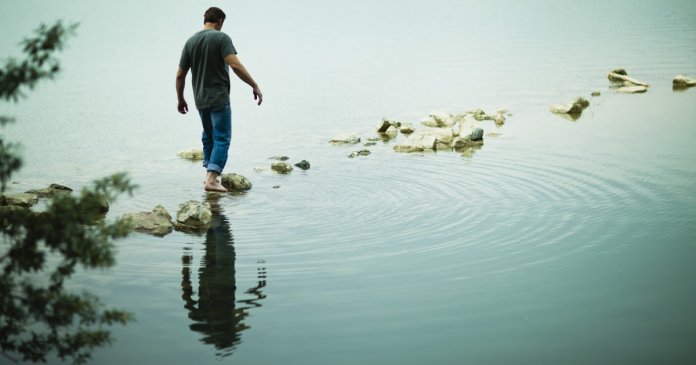 A man walking barefoot across stepping stones away from the shore of a lake.