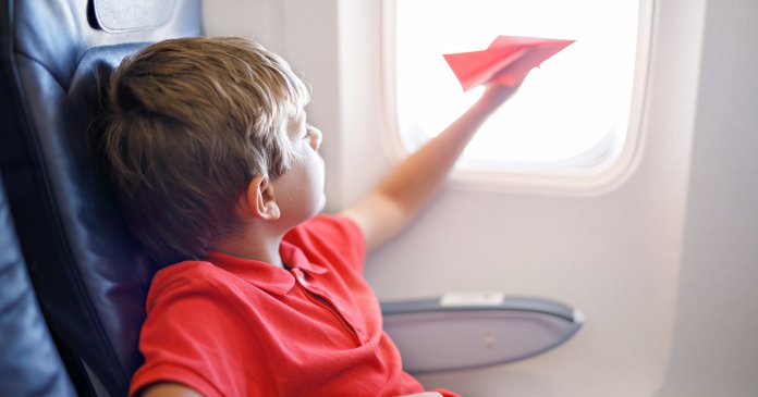 Little kid boy playing with red paper plane during flight on airplane