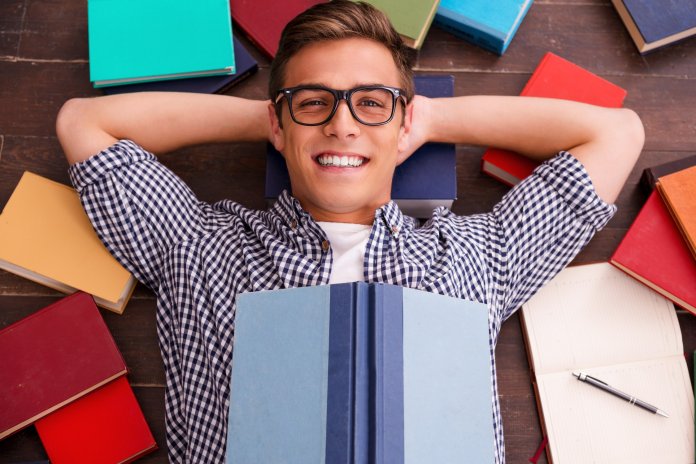 Reading is my hobby! Top view of happy young man holding hands behind head and smiling while lying on the hardwood floor with colorful books laying all around him